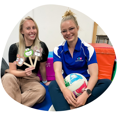 Lucy (SP, she/her) is pictured on the left and Monique (Physio, she/her) on the right. Lucy is holding up articulation visual resources and Monique is holding a netball, both are sitting around colourful soft play gym blocks and smiling at the camera
